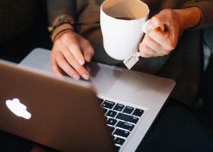 A woman's hands holding a mug of tea and an open laptop computer.