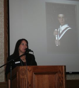 woman speaking into microphone at podium with young man's graduation photo projected behind her