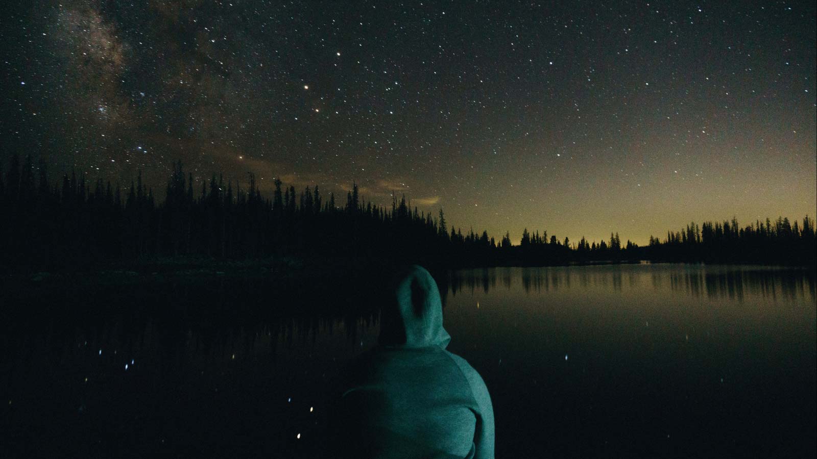 Person wearing hoodie sweatshirt looking out over a lake under starlight