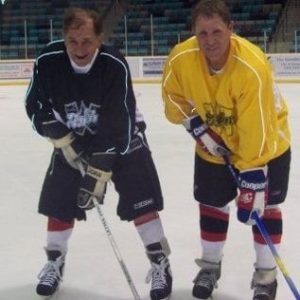 Two older men in hockey gear stand with sticks on the ice