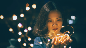 A young woman's face is lit by a strand of holiday lights she's holding in her cupped hands