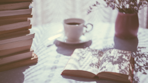 Filtered sunlight cast over a table with an open book, a stack of books, a plant, and a cup and saucer holding dark liquid