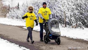 Man and woman wearing yellow T-shirts push stroller along snowy walkway. Both are wearing bright yellow T-shirts overtop of their jackets.