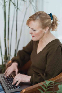 mature woman sits facing an open laptop. Her hands are positioned to type.