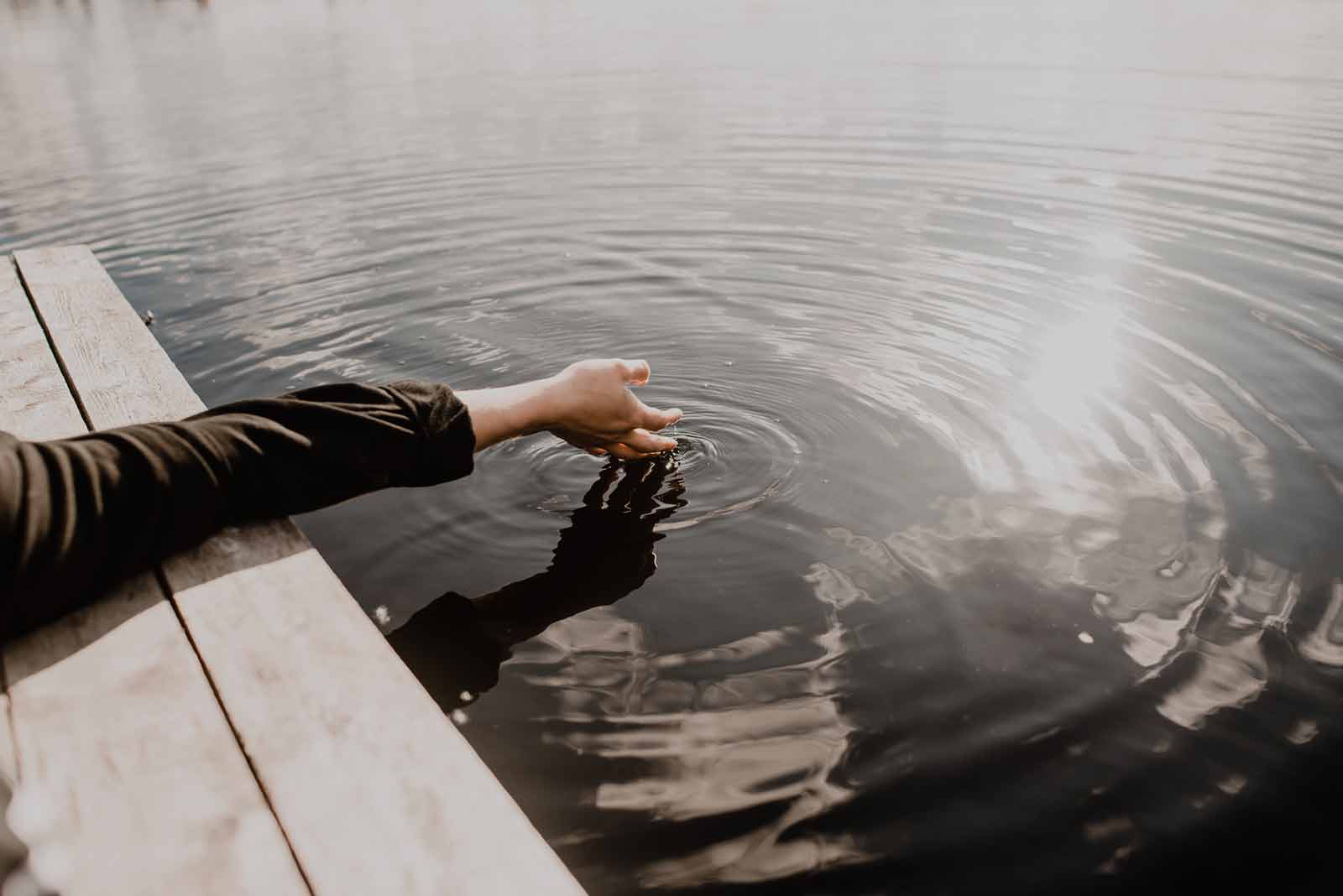 A black sleeved arm hangs into the water off of a dock. Ripples appear in the still water beneath the hand.