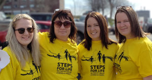 Four women standing shoulder to shoulder smile for the camera. They're each wearing a yellow T-shirt with a logo of a family that reads "Steps for Life"