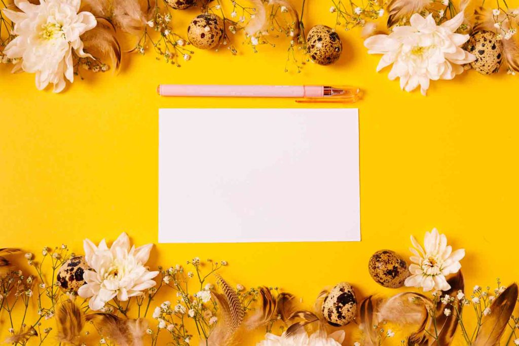 A blank piece of paper and a pen rest on a bright yellow background, framed with beautiful flowers and decorative eggs
