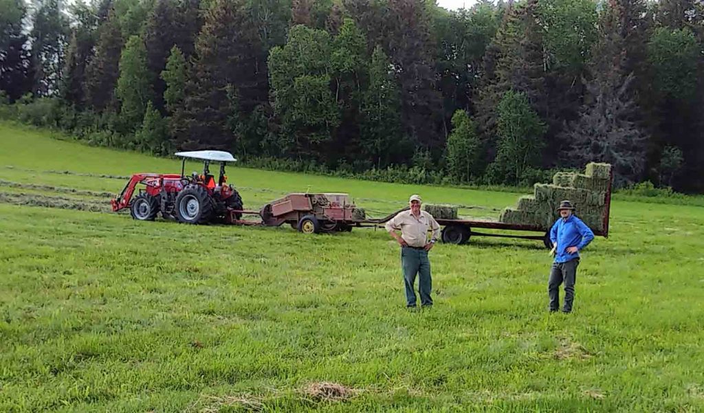 Two men stand in a field with a tractor and hay bales behind them