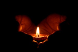 A pair of hands cup a lit candle. Surrounded by darkness, the glow of the light forms the shape of a heart.