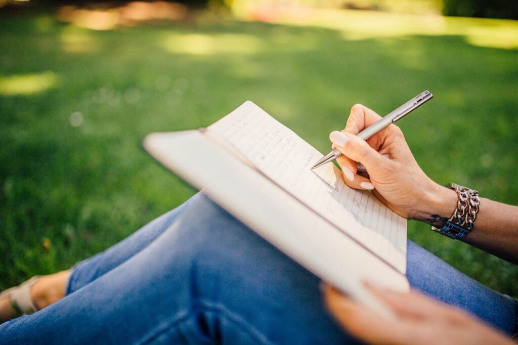 person sitting in the grass writing in a notebook