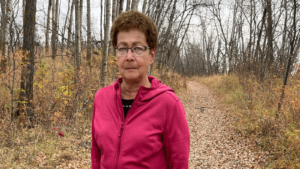 Woman with short brown curly hair and glasses stands stoically on a hiking path. She is dressed in a light hoodie jacket.