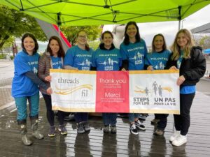 The Vancouver Steps for Life committee smile for a photo while holding the Steps for Life banner. The women are all wearing blue volunteer T-shirts.