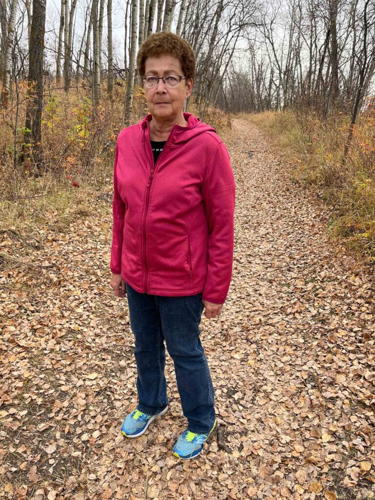Woman with short brown curly hair and glasses stands stoically on a hiking path. She is dressed in a light hoodie jacket. 