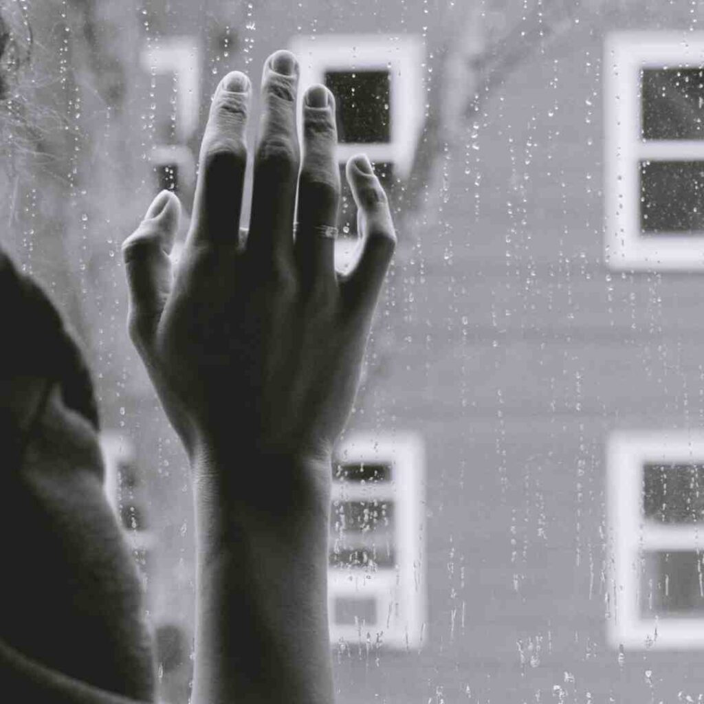 Black and white photo of a hand resting on a window; a building with rectangular windows is in the distance