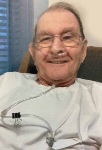 An older man wearing glasses smiles. He has an oxygen tube in his nose and draped down his chest.