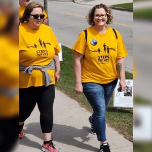 Melanie and a friend walk in Steps for Life t-shirts. Melanie is smiling brightly and wearing a button on her T-shirt.