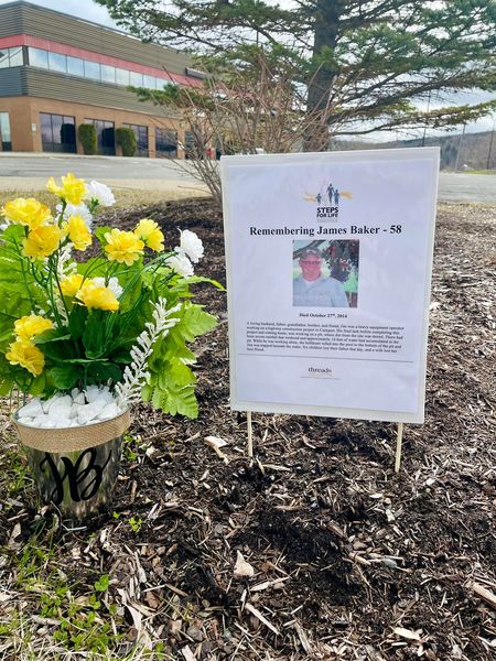 A sign titled "Remembering James Baker - 58" rests in the ground beside a potted plant of yellow and white flowers. The explanatory text on the sign is too small to read, but there is an image of a man wearing a blue long-sleeved shirt and a cap.