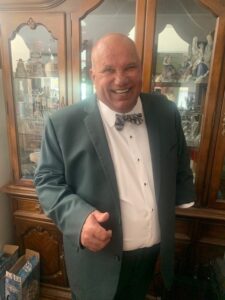 A smiling man, dressed in a suit, stands in front of a china cabinet. 