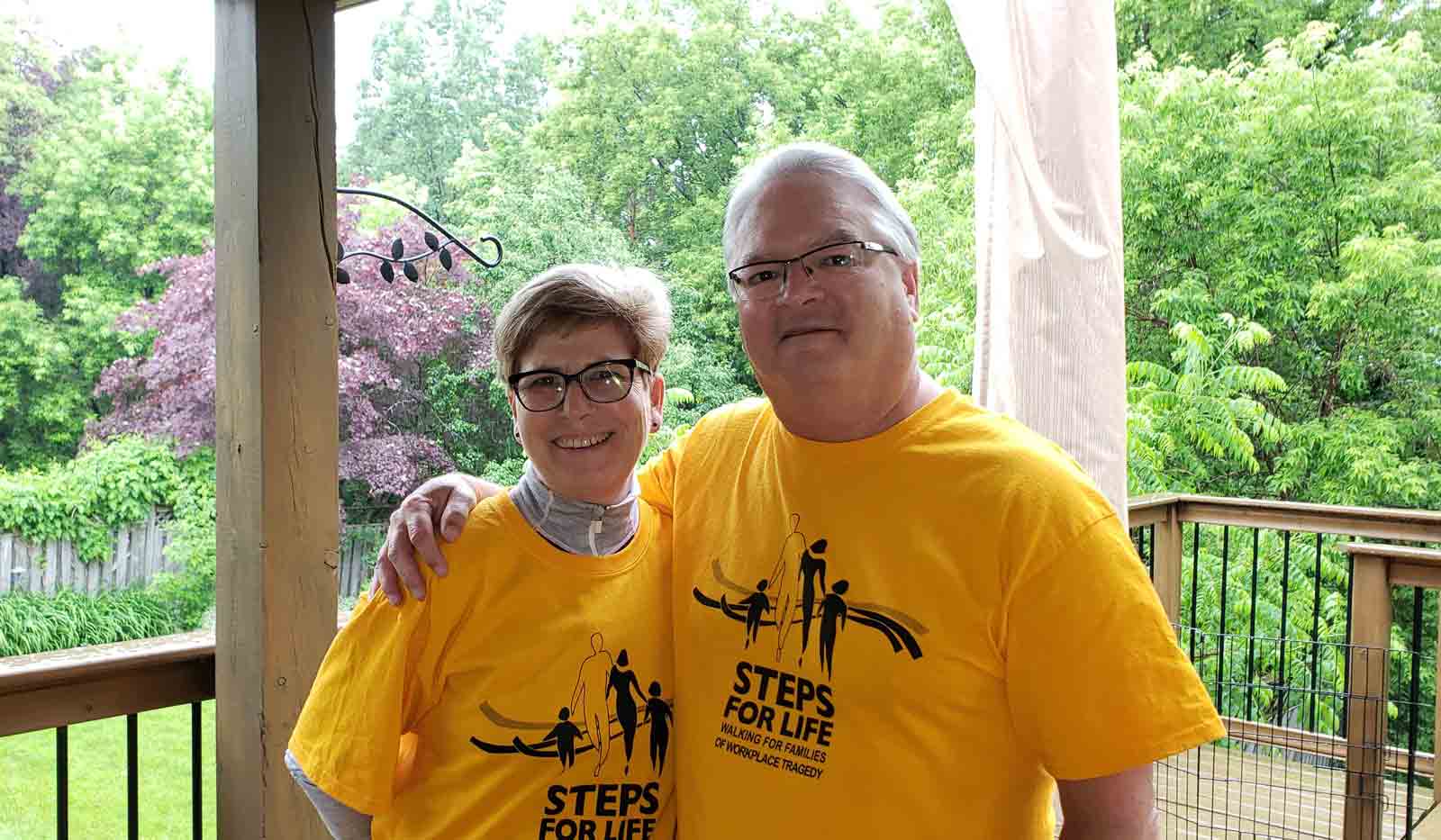 Marg and Scott pose in their yellow Steps for Life T-shirts. Lush greenery is seen behind them as they stand on the porch.
