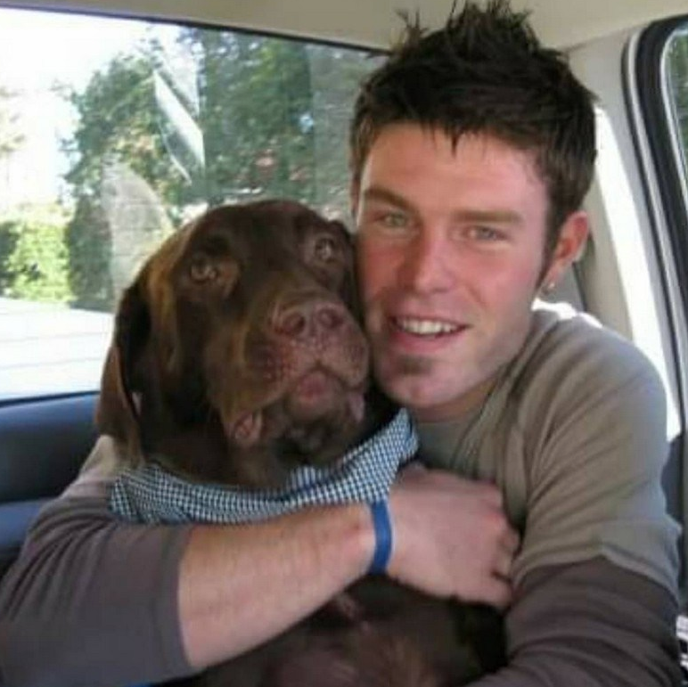 A man smiles while hugging his dog. They are seated in the cab of a vehicle. 