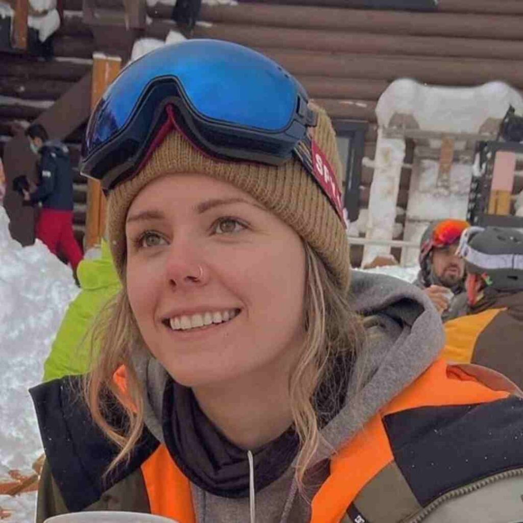 A young woman outfitted in ski goggles, toque, and coat smiles. She has blonde hair, fair skin, and is holding a beer in her hand.