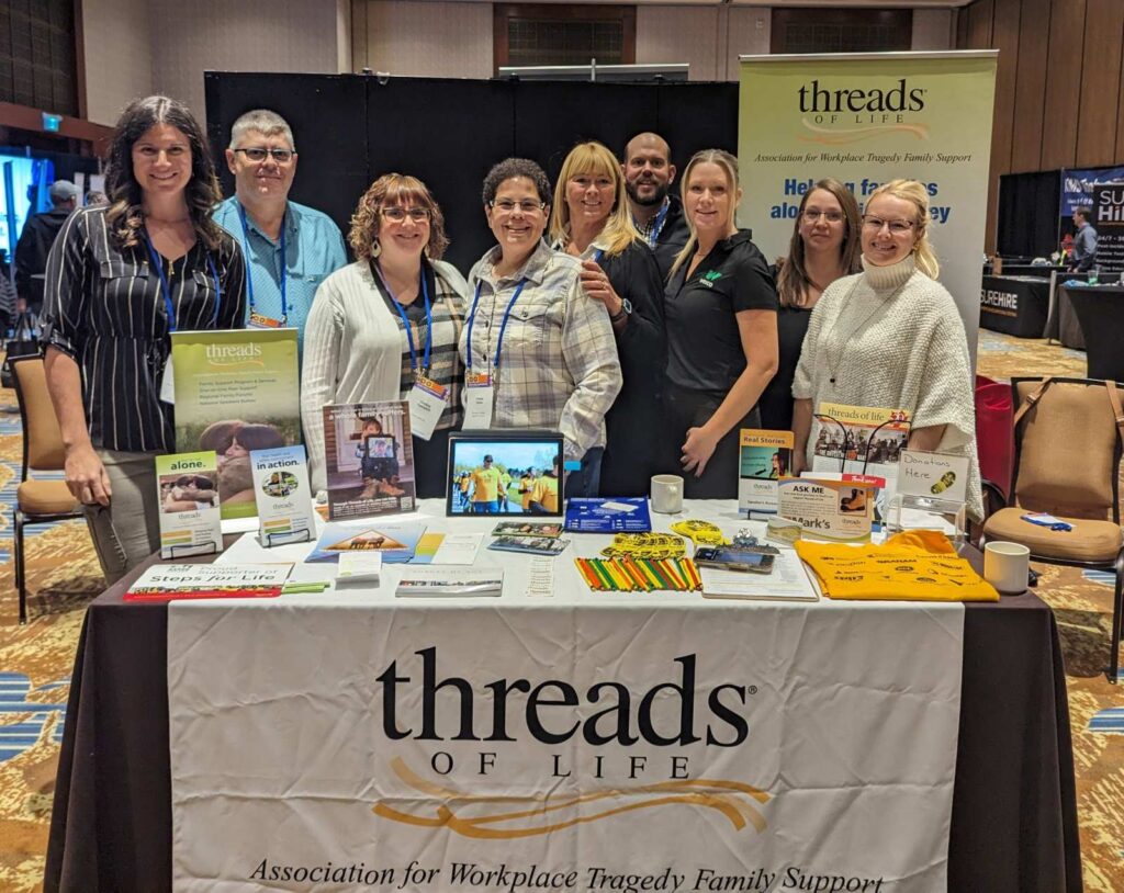 9 wonderful volunteers stand for a group photo behind a trade show booth displaying the Threads of Life logo and various materials. 