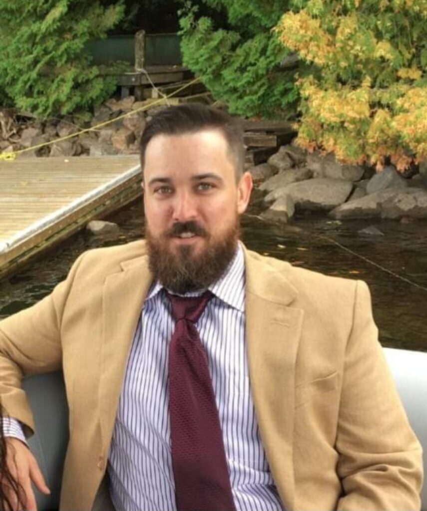 A man with a light complexion, brown hair and beard sits in a boat. He's wearing a tan blazer, striped shit and a maroon tie. The water is behind him and the dock, rocks and some cedar trees are visible on the shoreline.