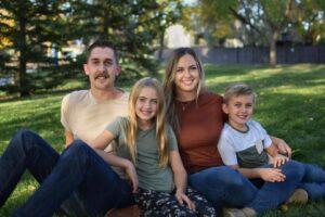 A man, woman and two young children - a girl and a boy - pose for a cozy family photo on a lawn.