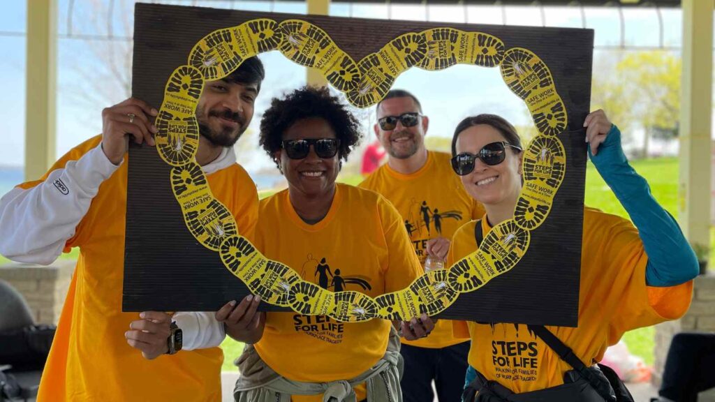 a group of 4 Steps for Life walkers smile as they hold a giant heart-shaped frame up around their faces. The line of the heart is framed with yellow Steps for Life bootprint images.