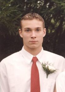 A young man stands facing the camera. He's wearing a dress shirt and a red tie.