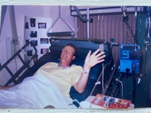 A man lying in a hospital bed raises his hand to wave.