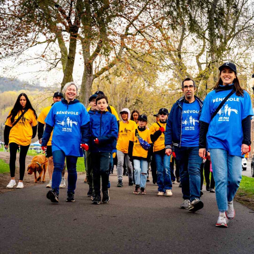 A group of walkers in matching T-shirts is walking toward the camera. The ones in front are volunteers wearing blue shirts that read "bénévole". The walkers behind them are participants wearing yellow Steps for Life t-shirts.