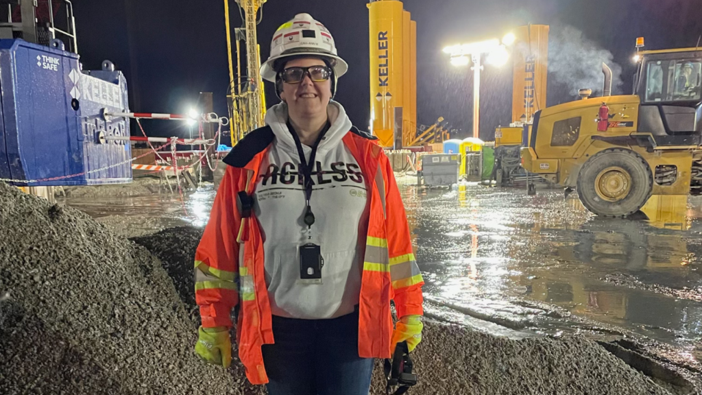 A woman in full safety gear stands on an industrial site. 